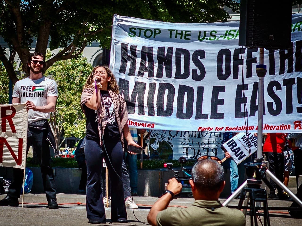 Speaker holding a microphone on a sunny day at a protest, sign in the background stays "hands off the Middle East"